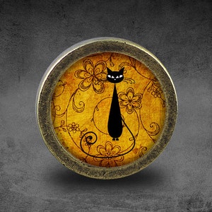 Könnte beinhalten: Ein runder, goldfarbener Metallrahmen mit einem Glaseinsatz, der eine schwarze Silhouette einer Katze mit einem floralen Muster auf gelbem Hintergrund zeigt.