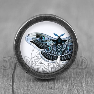 Könnte beinhalten: Ein silberner Knopf mit einer runden, weißen Fläche, die eine detaillierte Illustration eines blau-schwarzen Schmetterlings mit blauen Augen zeigt. Der Schmetterling ist von einem weißen Blumenmuster umgeben.