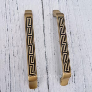 Pode incluir: Duas puxadores de armário de latão antigo com um design geométrico preto. Os puxadores têm formato retangular com uma alça curva e são colocados sobre um fundo de madeira branca. Os puxadores têm aproximadamente 15 cm de comprimento.