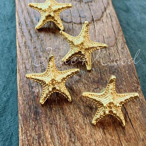 Op de afbeelding: Vier gouden, stervormige kastknoppen op een houten oppervlak. De sterren zijn getextureerd en hebben een licht verhoogd ontwerp.