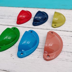 Puede incluir: Seis pomos de armario de media luna de colores, en varios colores, incluyendo rojo, azul, verde, amarillo, azul claro y naranja. Los pomos están dispuestos en una fila sobre una superficie blanca.