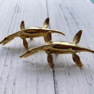 Puede incluir: Dos figuritas de dinosaurio de color dorado, posiblemente de latón, se muestran sobre una superficie de madera blanca. Los dinosaurios están mirando en direcciones opuestas, con sus colas enroscadas.