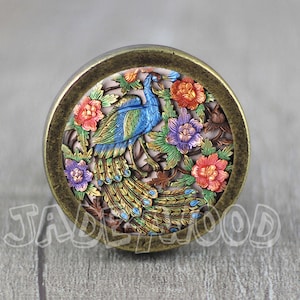 May include: A round, bronze-colored knob with a detailed peacock design. The peacock is blue, green, and gold with a long tail. The background is a light pink with floral accents.