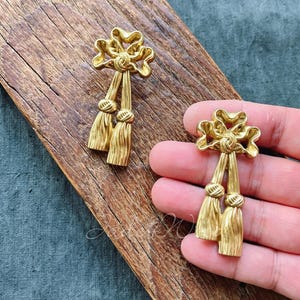 Op de afbeelding: Een paar gouden oorbellen met een strik- en kwastontwerp. De oorbellen zijn gemaakt van metaal en hebben een glanzende afwerking.