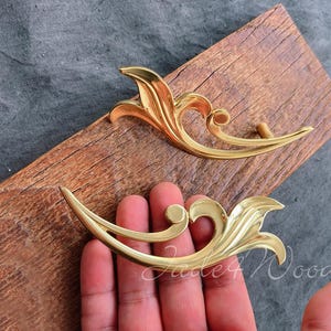 Puede incluir: Dos tiradores de cajones decorativos de color dorado con diseños ornamentados. Los tiradores son de metal y tienen un acabado brillante.
