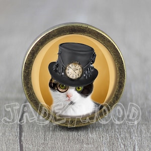 May include: A circular button with a bronze-effect frame, showcasing a black and white cat in a steampunk-style top hat with a clock face and a monocle. The cat has green eyes, set against a yellow backdrop. Text reads "JADEWOOD".