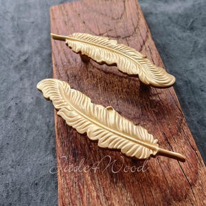 Pode incluir: Duas puxadores de armário em forma de pena douradas em uma superfície de madeira. Os puxadores têm um design detalhado e texturizado.