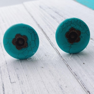 May include: Two turquoise blue earrings with a gold flower design. The earrings are round and have a textured surface.