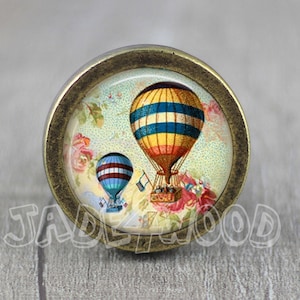 Puede incluir: Un pomo de armario de estilo vintage con una base redonda de color latón y una parte superior de vidrio con una ilustración colorida de dos globos aerostáticos sobre un fondo floral.