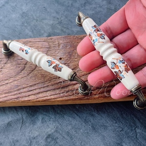 Peut inclure: Deux boutons de cabinet en céramique blanche avec un motif floral dans des tons d'orange, de bleu et de brun. Les boutons ont une finition en laiton brossé et sont montés sur une surface en bois.