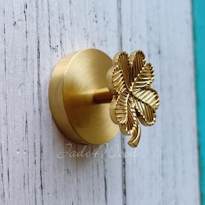 May include: A brass-coloured cabinet knob featuring a four-leaf clover design. The clover is intricately detailed and attached to a circular base. The knob is mounted on a white wooden surface. The text "Jade4Wood" is visible.