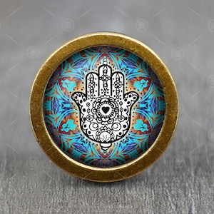Peut inclure: Un bouton rond doré avec un motif de mandala bleu, turquoise et orange représentant une main de hamsa blanche avec un contour noir et un petit cœur au centre.