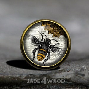 Może przedstawiać: Okrągły, złoty guzik z szklanym wierzchem przedstawiający ilustrację czarnej i żółtej pszczoły na tle strony z antycznego tomu. Pszczoła leci w pobliżu plastra miodu.