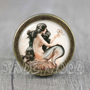 Puede incluir: Pomo de armario redondo, tono latón antiguo, con una ilustración vintage de una sirena. La sirena tiene pelo largo y negro y una cola de pez. Sostiene un zapato. El texto "JADEWOOD" es visible.