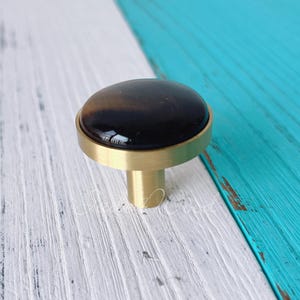 May include: A cabinet knob with a dark, oval-shaped stone set in a gold-colored metal base. The knob has a cylindrical stem and is set against a white and turquoise painted wooden surface.