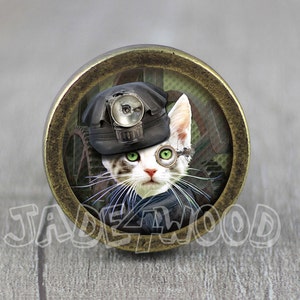 May include: A white and grey cat wearing a black miner's hat with a headlamp and a round, green eye patch. The cat is looking directly at the camera. The image is in a round, brass-coloured frame.