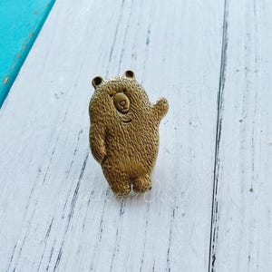 Puede incluir: Una figurita de oso de color dorado con una superficie texturizada. El oso está de pie sobre una superficie de madera blanca.