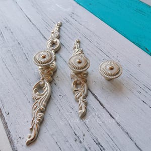 May include: Three ornate silver drawer pulls with a gold accent. The pulls have a floral design and are arranged on a white wooden surface.