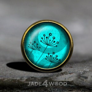 May include: A turquoise blue glass cabochon with a brass setting features a stylised design of three black dandelions. The cabochon is set against a textured background.