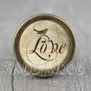 May include: A round, antique gold-toned knob with a vintage-style image of a bird perched on a branch with the word "Love" in a decorative script.