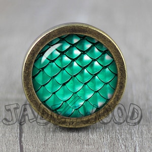 Könnte beinhalten: Ein runder, grüner und bronzefarbener Dekorknopf mit einem Schuppenmuster. Der Knopf ist aus Metall und Glas.
