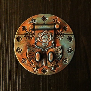 Puede incluir: Un cierre de metal circular adornado con un diseño de mariposa. El cierre está decorado con una rosa, flores y espirales. El cierre es de color naranja oxidado con detalles azules.