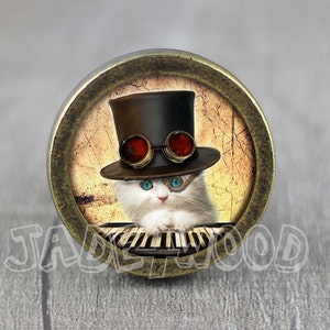 May include: A circular button with a bronze-coloured rim, showcasing a white cat in a top hat and steampunk goggles. The cat is positioned in front of a piano keyboard. The background has a vintage, textured look. The text "JADEWOOD" is visible.