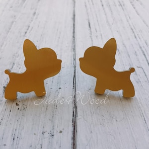 May include: Two gold-colored French bulldog-shaped earrings. The earrings are mirror images of each other, each with a rounded body, small tail, and large, upright ears. The earrings are set against a white and light blue wooden background.