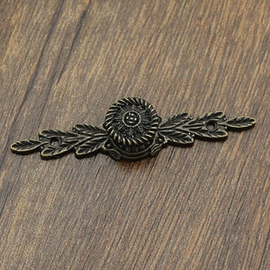 Puede incluir: Un adorno de metal decorativo con un diseño floral. El adorno es de color bronce oscuro y tiene un centro circular con un diseño en relieve. El adorno es perfecto para agregar un toque de encanto vintage a cualquier proyecto.