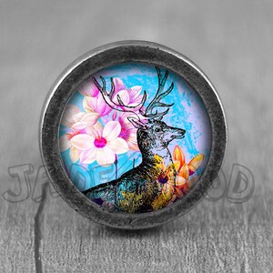 Op de afbeelding: Een ronde, zilverkleurige knop met een kleurrijk beeld van een hert met geweien dat in een veld met roze en gele bloemen staat. De achtergrond is een lichtblauwe aquarelwas.