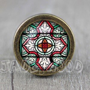 Könnte beinhalten: Ein runder, messingfarbener Schrankknopf mit einem Buntglasdesign. Das Buntglas zeigt ein rotes, grünes und weißes Muster mit einem zentralen Kreuz.