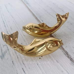 Könnte beinhalten: Zwei goldfarbene Fischfiguren. Die Fische sind gebogen, mit angehobenen Schwänzen und Flossen. Die Figuren haben detaillierte Schuppen und Augenmerkmale. Der Hintergrund ist eine helle, strukturierte Oberfläche.