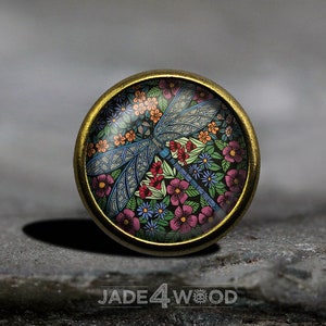 Peut inclure: Un bouton rond en métal doré avec un dôme en verre représentant une libellule colorée entourée de fleurs. La libellule est bleue, noire et blanche, et les fleurs sont roses, rouges, orange, jaunes et bleues.