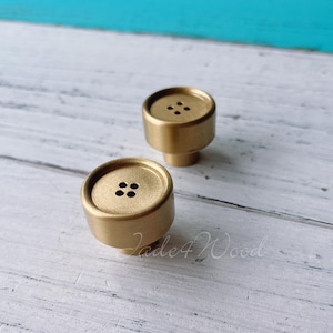 May include: Two gold-toned metal cabinet knobs with a round button design and four small holes in the center. The knobs are on a white wooden surface.