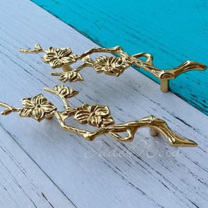 Op de afbeelding: Twee gouden kastknoppen in de vorm van gestileerde takken met delicate bloemdetails. De knoppen zijn ontworpen om te lijken op een bloeiende tak.