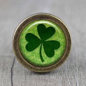 Könnte beinhalten: Ein runder, messingfarbener Schrankknopf mit einem grünen Kleeblatt-Design. Das Kleeblatt ist von einem grünen Hintergrund mit kleineren Kleeblättern umgeben.