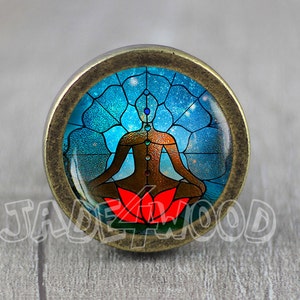 May include: A round, brass-coloured cabinet knob with a glass insert featuring a colourful stained glass design of a person meditating in a lotus position. The person is surrounded by a blue and purple stained glass background with a star-like pattern.