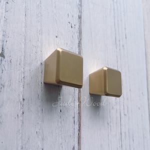 May include: Two square, gold-colored cabinet knobs mounted on a white, distressed wooden surface. The knobs are cube-shaped with rounded edges. The text "Jade + Wood" is written in cursive on the surface.