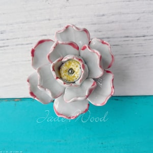 May include: A ceramic flower-shaped cabinet knob with gray petals edged in pink. The center is yellow with a silver accent. The knob is set against a white and turquoise background. The text "Jade Wood" is visible.