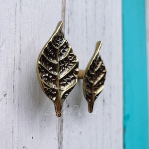 Könnte beinhalten: Zwei messingfarbene Schrankgriffe in Blattform. Die Griffe sind mit Adern versehen und haben eine warme, antike Oberfläche. Sie sind auf einer weiß und türkis lackierten Holzoberfläche montiert. Sie verleihen Möbeln eine natürliche, elegante Note.