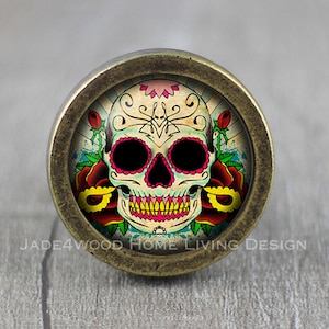 May include: A round, antique gold-toned cabinet knob with a colourful sugar skull design. The skull is decorated with pink, yellow, and red flowers.
