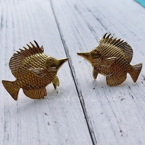 May include: A pair of gold-tone fish-shaped earrings. The fish have a detailed, textured design and are facing opposite directions.