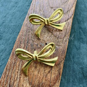 Può includere: Due pomelli per armadi a forma di fiocco di colore dorato. I pomelli sono su una superficie di legno.