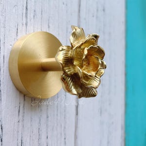 Peut inclure: Un crochet mural doré en forme de fleur. Le crochet présente un motif floral détaillé en trois dimensions avec de multiples pétales. Le crochet est monté sur une base circulaire et est fait d'un matériau métallique brillant. Le texte "Jade4Wood" est visible.