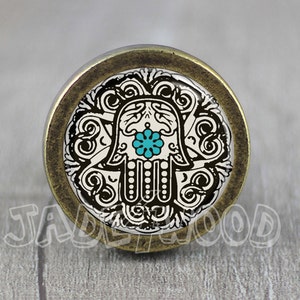 Peut inclure: Un bouton de porte rond en bronze avec un motif décoratif. Le motif représente une main de hamsa noire et blanche avec un œil bleu turquoise au centre. La main de hamsa est entourée de motifs floraux complexes.