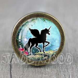 May include: A round, brass-colored cabinet knob with a glass insert featuring a black silhouette of a unicorn with wings in a blue and green forest scene.