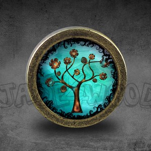 May include: A round, brass-coloured knob with a decorative glass insert. The glass features a stylised tree with brown branches and orange flowers on a teal background. The tree is surrounded by a black, ornate border.