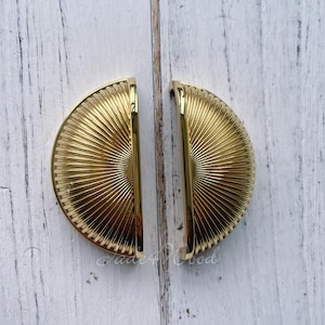 Może przedstawiać: Dwa złote uchwyty do szafek z wzorem słońca. Uchwyty mają kształt półkola i gładkie, błyszczące wykończenie.