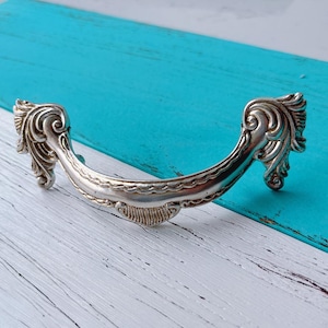 May include: Silver-toned decorative drawer pull with an ornate design. The pull is shaped like a crescent moon with a detailed, scroll-like pattern.