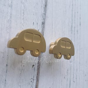 Pode incluir: Dois puxadores dourados em forma de carro. Cada puxador tem um corpo retangular arredondado com duas janelas retangulares e duas rodas circulares. Os puxadores são feitos de metal e são projetados para móveis. O fundo é uma superfície de madeira clara.